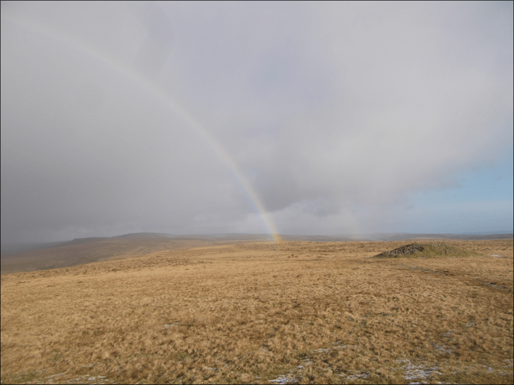 A view across Harford Moor on a cloudy day in winter - the grass is brown with some snow, the sky is mostly grey with a double rainbow slanting across it. To the right is an ancient barrow, a pile of earth and stone.