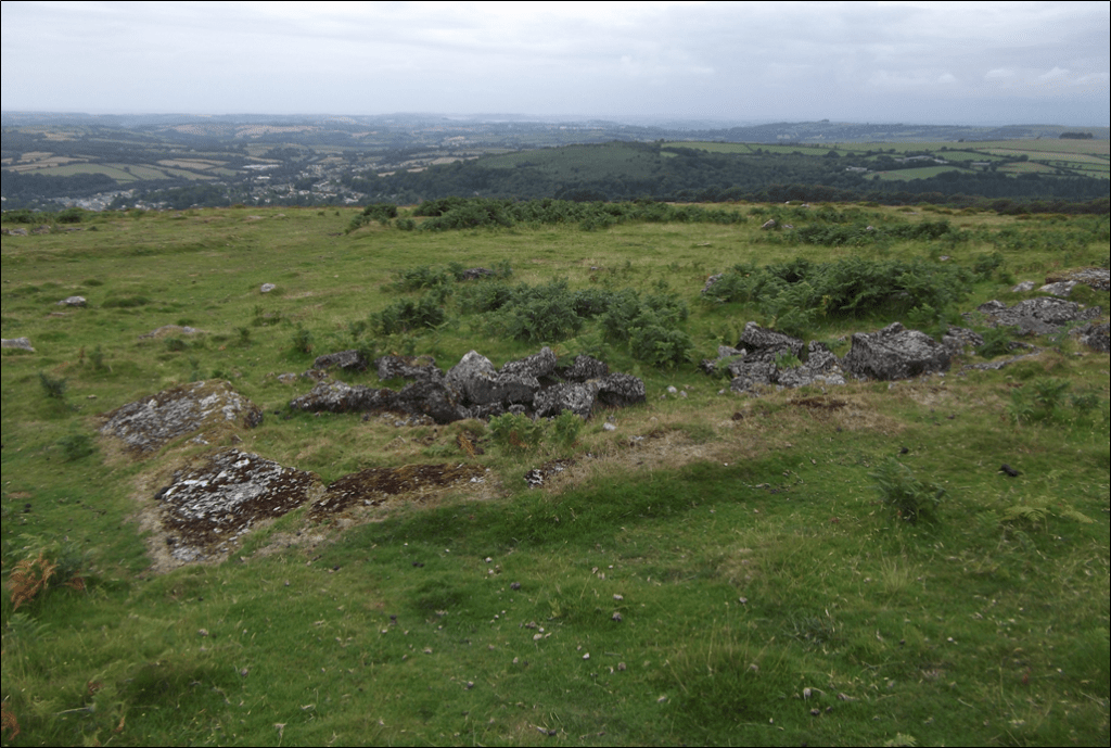 Concrete foundations and rubble among the grass and bracken on the Western Beacon, with Ivybridge and Henlake Down in the distance.